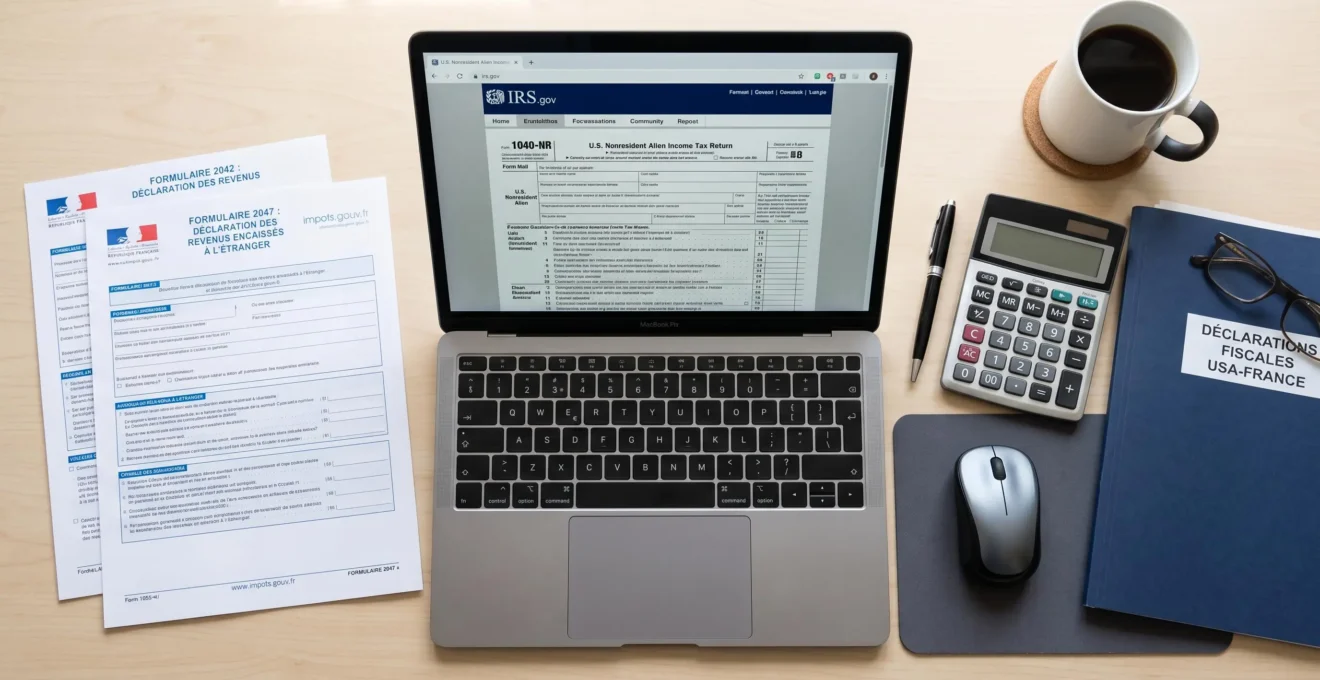 Bureau de travail organisé en vue de dessus avec ordinateur affichant le site IRS.gov (formulaire 1040-NR), documents fiscaux français imprimés (formulaire 2047), calculatrice et dossier de déclarations fiscales USA-France